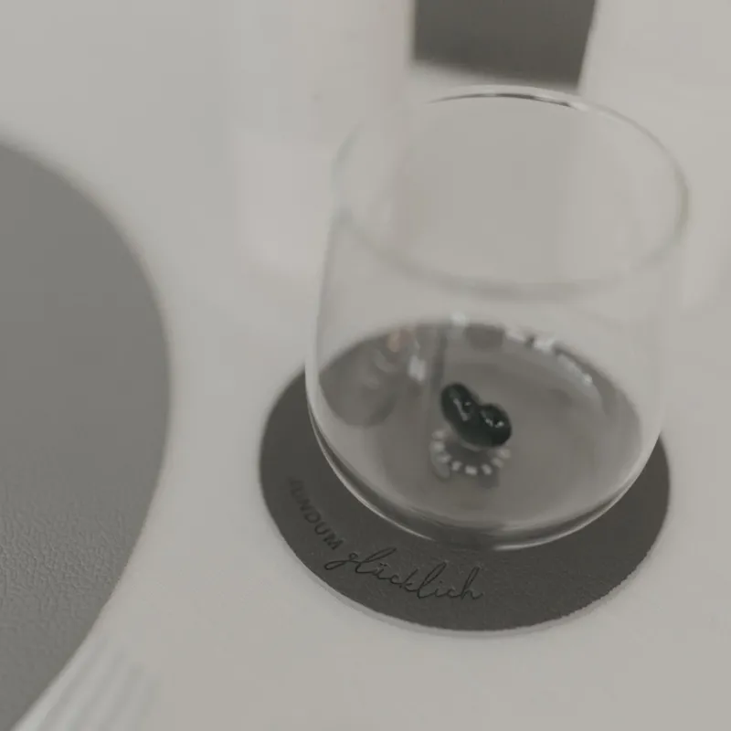 Grauer Untersetzer „Rundum glücklich“ mit Glas darauf, modernes Design auf hell gedecktem Tisch.